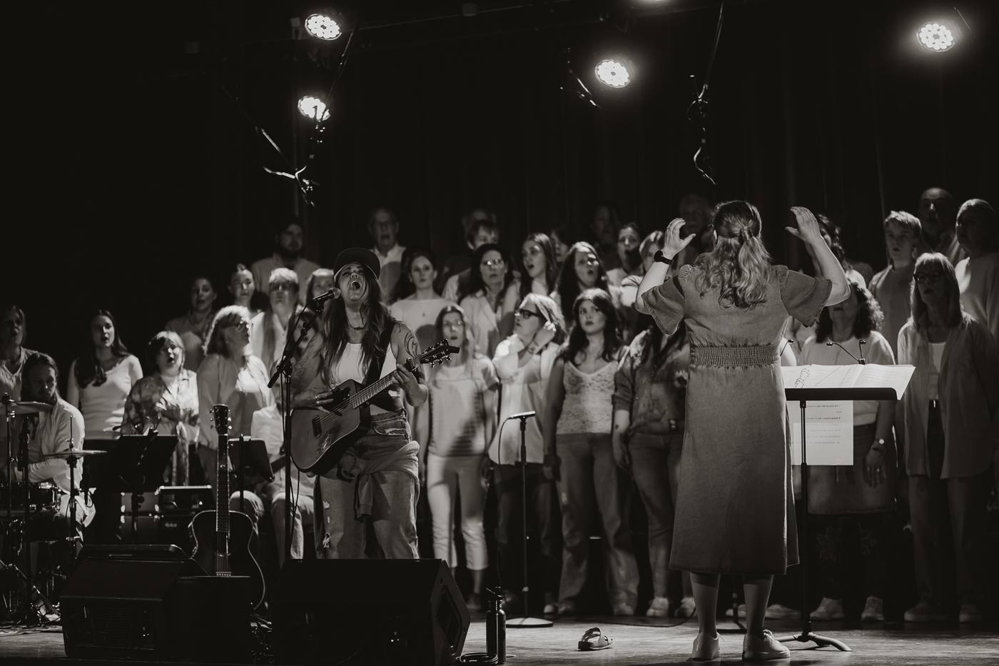 Deborah Edin sjunger med gitarr framför en stor projektkör på Röda Kvarn i Orsa, körledaren dirigerar i förgrunden.